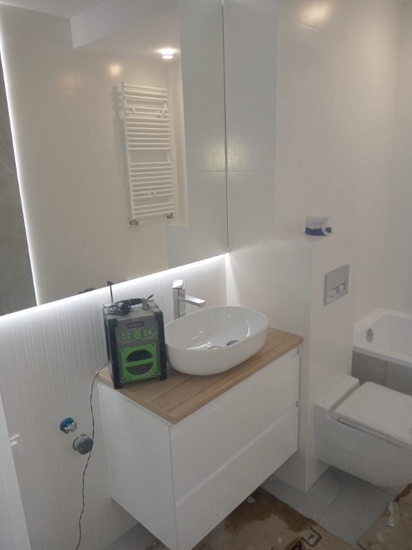 Nowoczesna, jasna łazienka z umywalką nablatową na drewnianym blacie, widoczny grzejnik drabinkowy i radio budowlane obok umywalki.