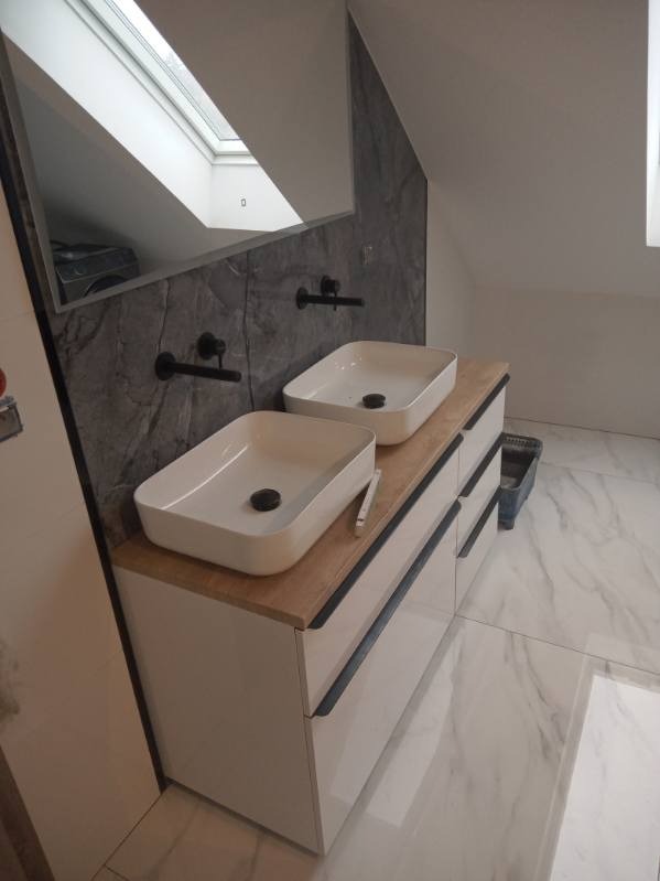 Dwie białe, prostokątne umywalki nablatowe z czarną armaturą na drewnianym blacie szafki w łazience z marmurowymi płytkami i oknem dachowym widocznym w lustrze.