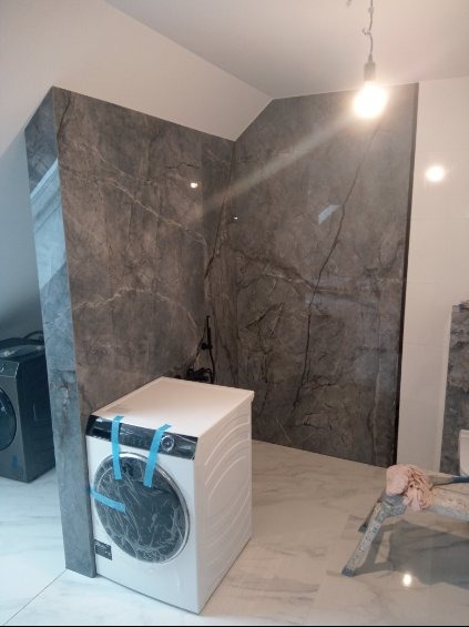 Nowa pralka, zabezpieczona taśmą, stoi w rogu wykończonej łazienki z szarymi płytkami imitującymi marmur na ścianach i podłodze.