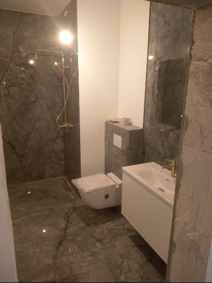 Wykończona łazienka z szarymi płytkami imitującymi marmur, złotą armaturą prysznicową i umywalkową, białą szafką pod umywalką i podwieszaną toaletą z zakrytą spłuczką. Na blacie obudowy toalety...