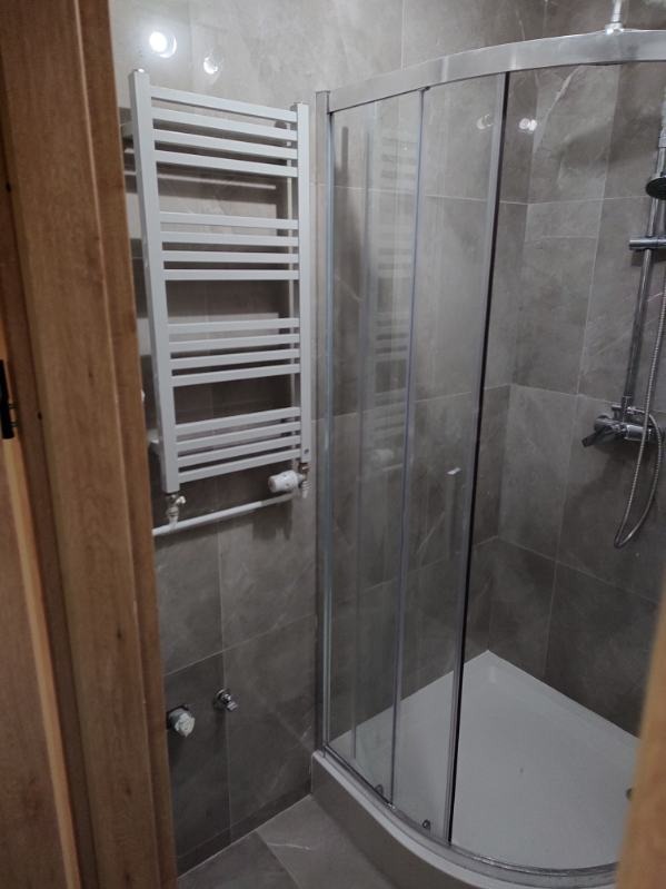 Szara łazienka z kabiną prysznicową z zaokrąglonymi drzwiami i grzejnikiem drabinkowym na ścianie z płytek.