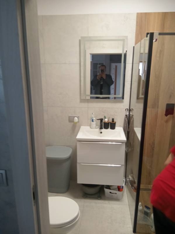 Wykończona łazienka z beżowymi płytkami, białą szafką pod umywalką, toaletą z szarą obudową i kabiną prysznicową z drewnopodobnymi panelami, widoczny refleks fotografa w lustrze.