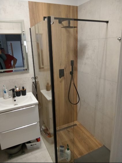 Nowoczesna łazienka z kabiną prysznicową typu walk-in, wykończoną płytkami imitującymi drewno, czarną armaturą i umywalką z szafką w kolorze białym; odbicie osoby w lustrze.