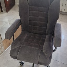 Usługi Tapicerskie Sebastian Kasperski - Szare, tapicerowane krzesło biurowe z podłokietnikami, wykończone pomarańczową nicią, na kółkach, stojące na płytkach w pomieszczeniu.