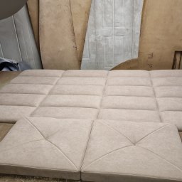 Usługi Tapicerskie Sebastian Kasperski - Panel tapicerowany w beżowym odcieniu, składający się z kwadratowych elementów z przeszyciami, leżący na drewnianym stole w warsztacie tapicerskim.