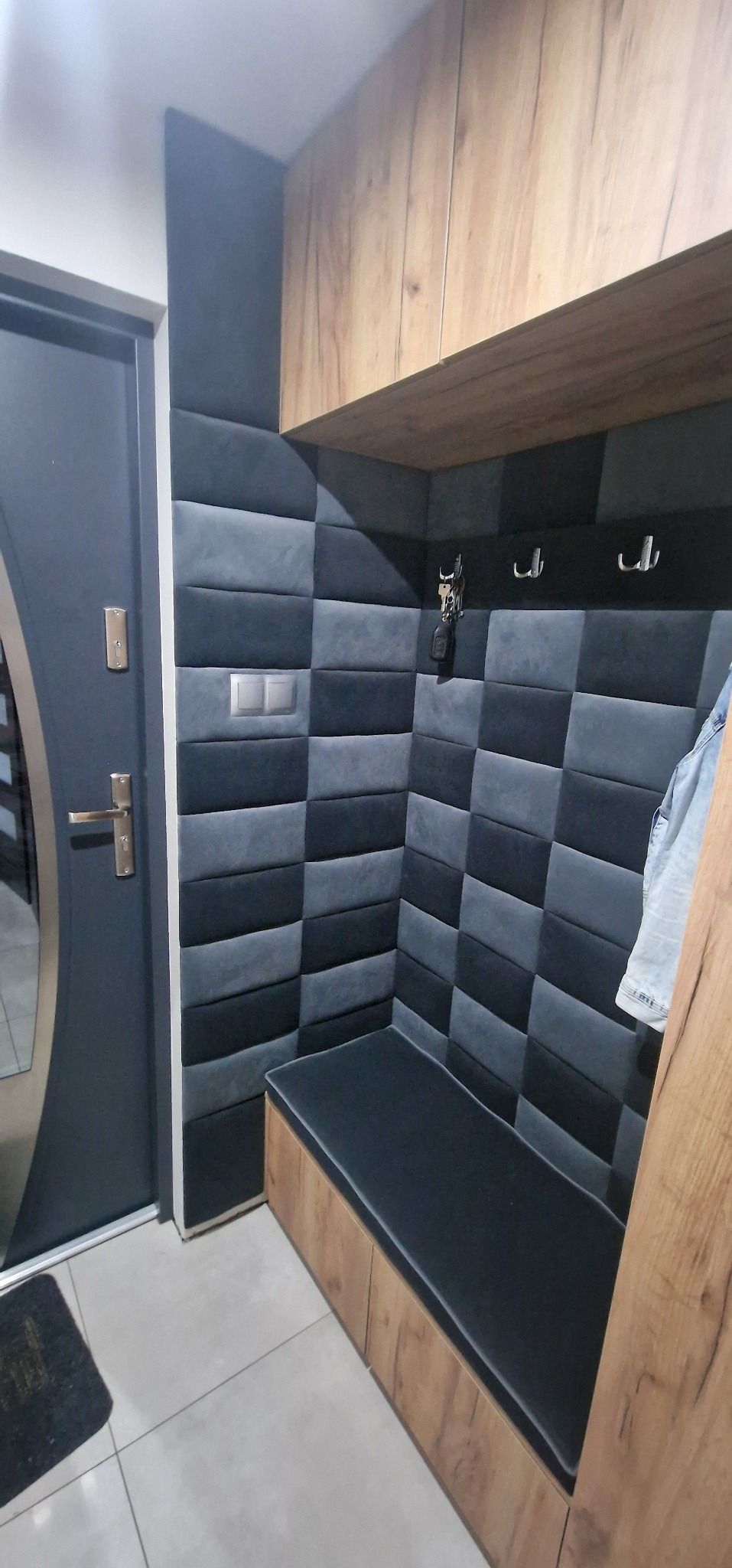 Tapicerowany panel ścienny w szaro-czarną szachownicę z wbudowaną tapicerowaną ławką w przedpokoju, obok drzwi wejściowych.