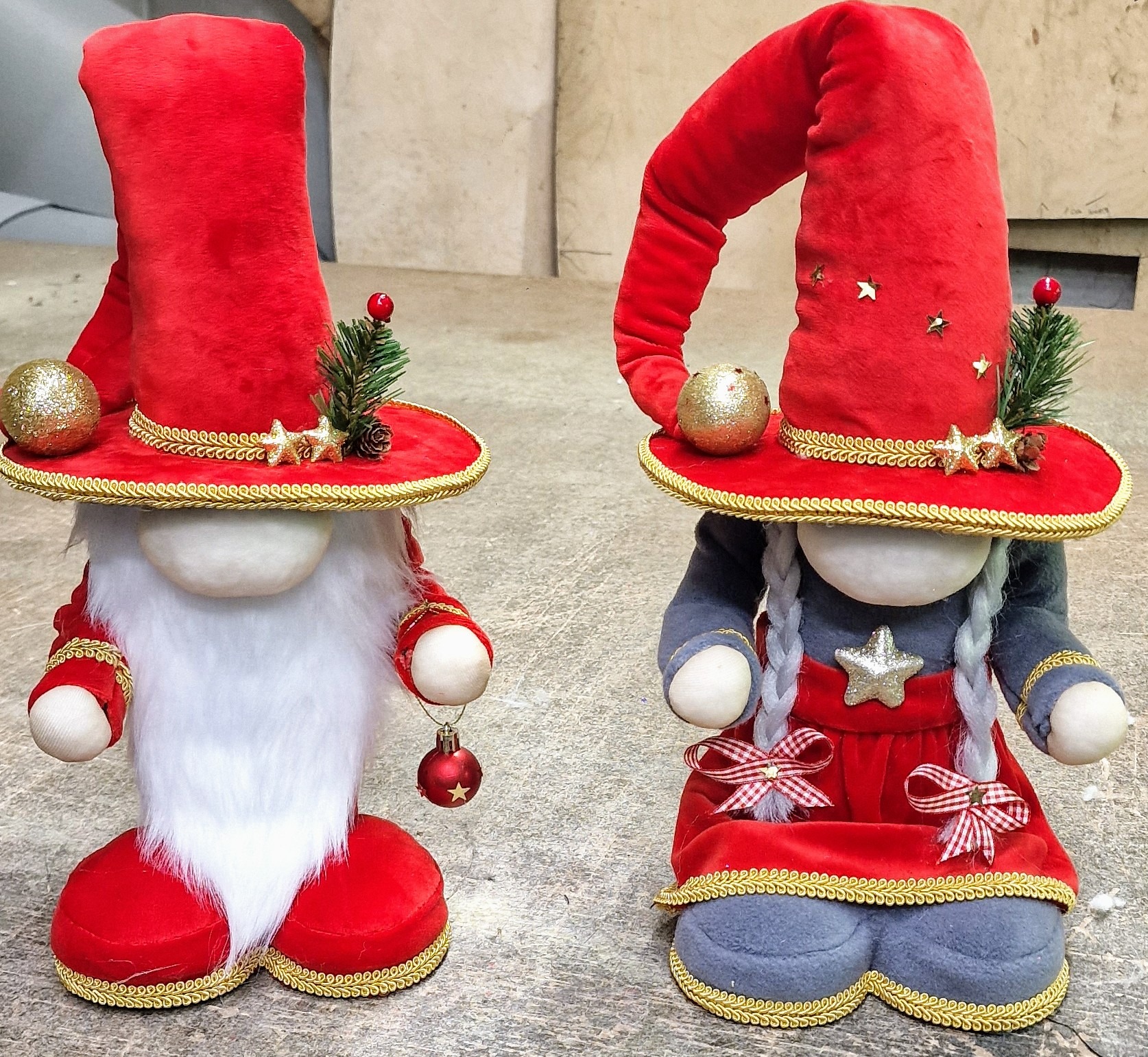 Dwa ręcznie robione, świąteczne gnomy z czerwonymi kapeluszami i złotymi zdobieniami. Jeden z białą brodą i czerwoną ozdobą, drugi z szarymi warkoczami i czerwoną sukienką.