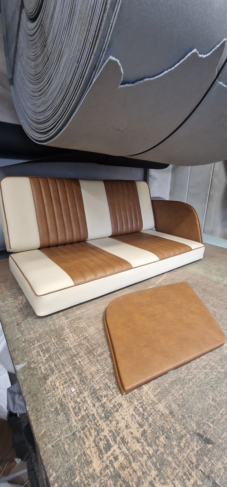 Kanapa z brązowo-kremową tapicerką w paski, obok leży poduszka, nad nią rolka materiału tapicerskiego