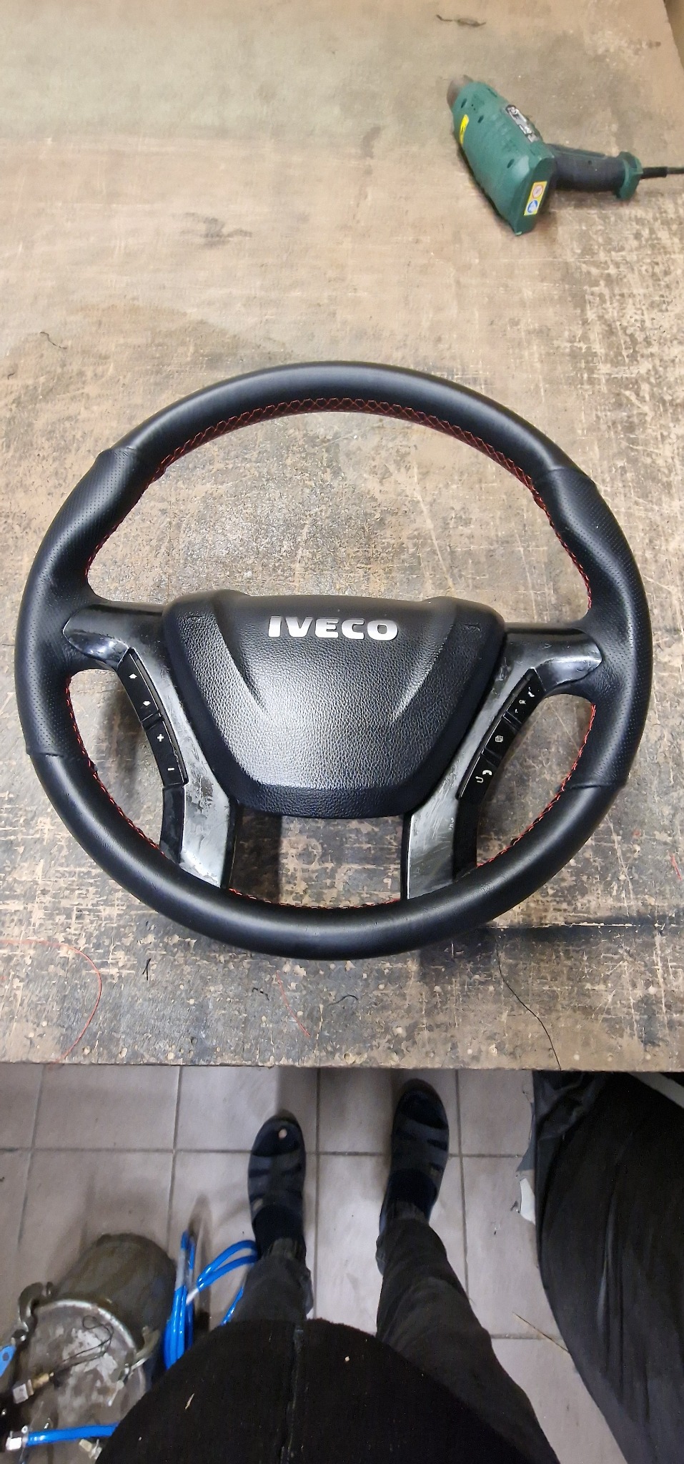 Odnowiona kierownica Iveco z czarną skórą i czerwonym szwem, leżąca na warsztatowym stole, obok opalarki.