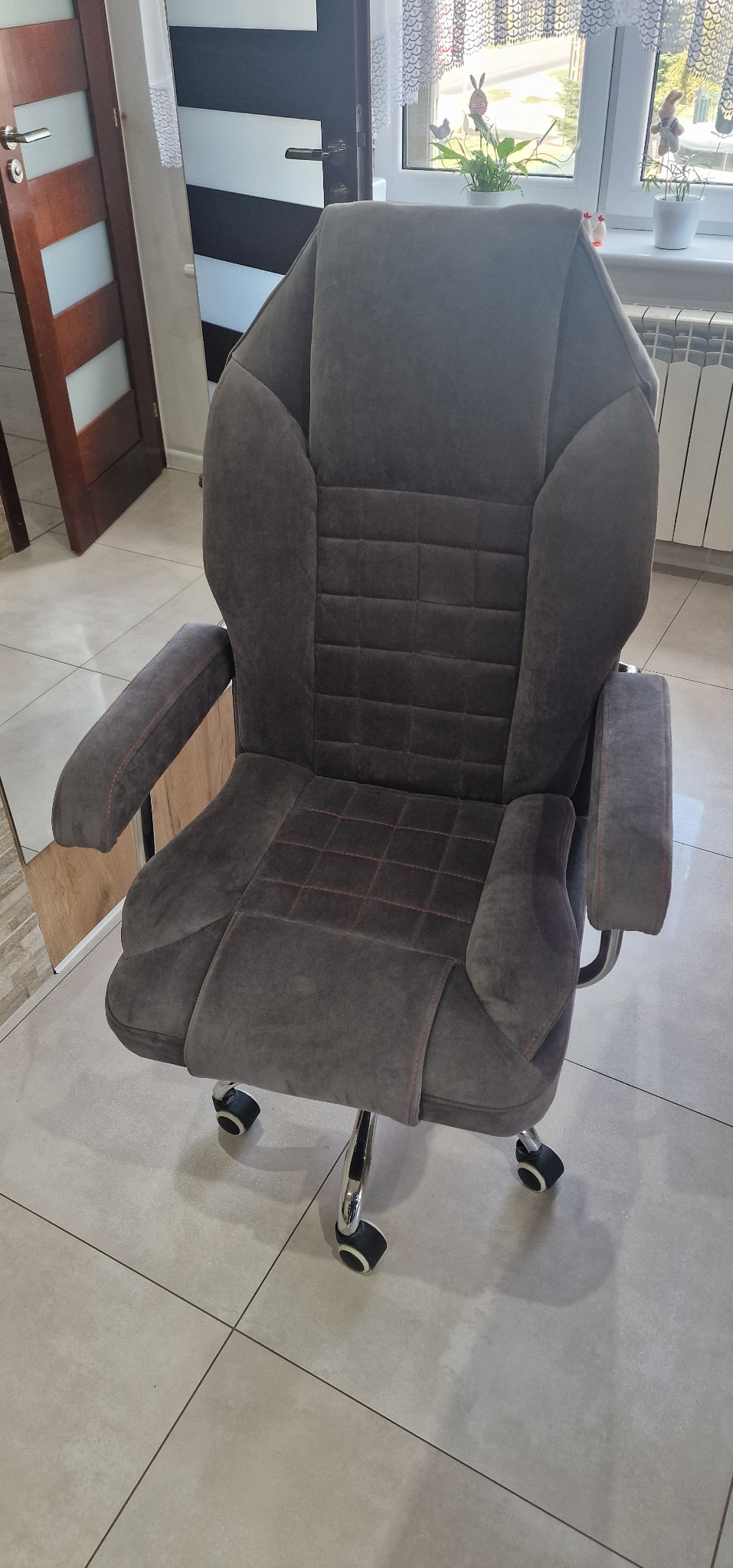 Szare, tapicerowane krzesło biurowe z podłokietnikami, wykończone pomarańczową nicią, na kółkach, stojące na płytkach w pomieszczeniu.