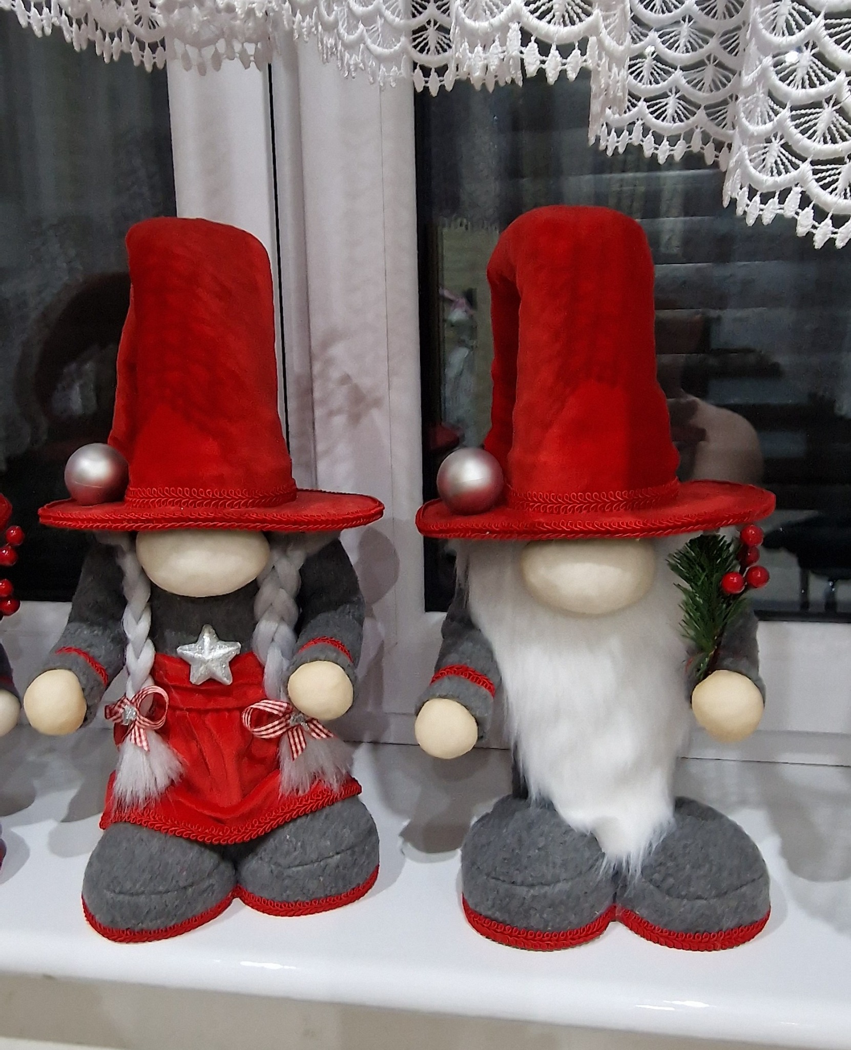 Dwa ozdobne, ręcznie robione skrzaty w czerwono-szarych strojach stojące na parapecie okiennym, jeden z warkoczami, drugi z brodą, obaj z czerwonymi kapeluszami i srebrnymi kulkami.