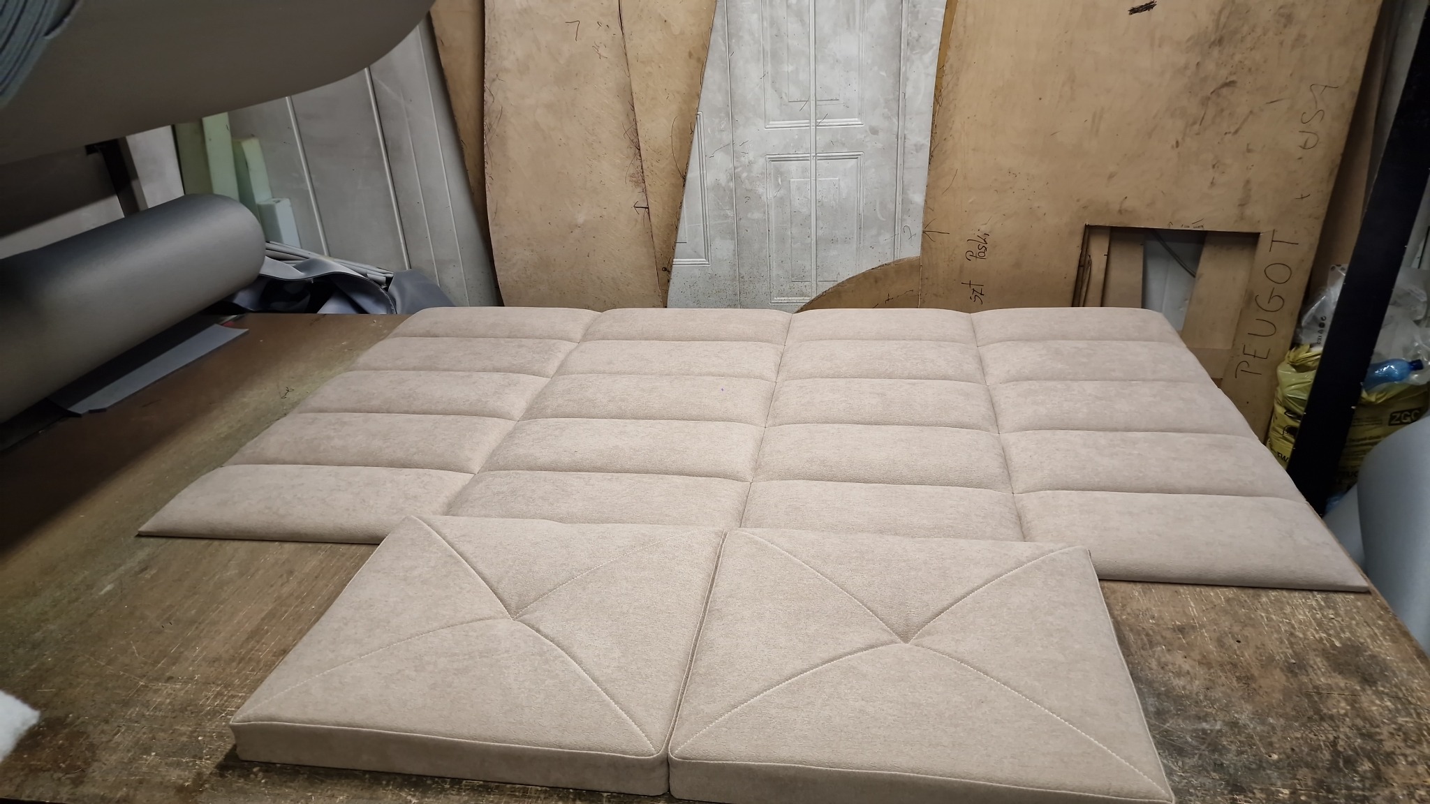 Panel tapicerowany w beżowym odcieniu, składający się z kwadratowych elementów z przeszyciami, leżący na drewnianym stole w warsztacie tapicerskim.