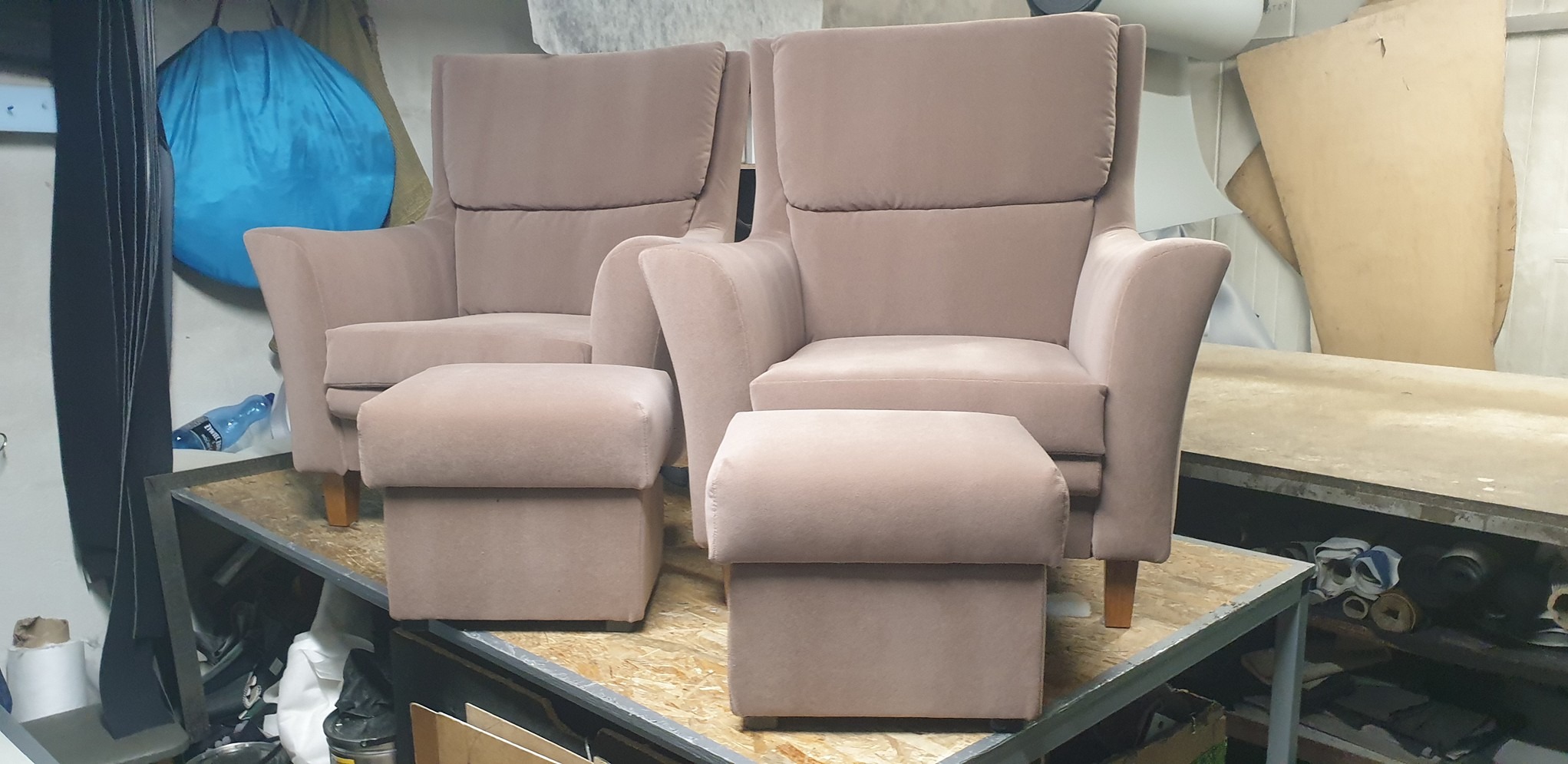 Dwa beżowe fotele tapicerowane z podnóżkami, ustawione na metalowej konstrukcji w warsztacie tapicerskim, w tle widoczne materiały i narzędzia.