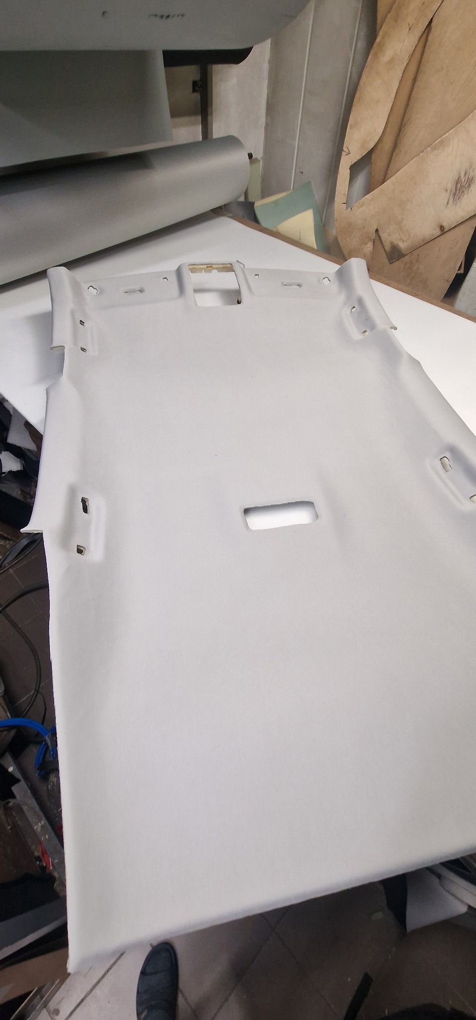 Jasnoszara podsufitka samochodowa z wycięciami na oświetlenie i mocowania, leżąca na stole w warsztacie.