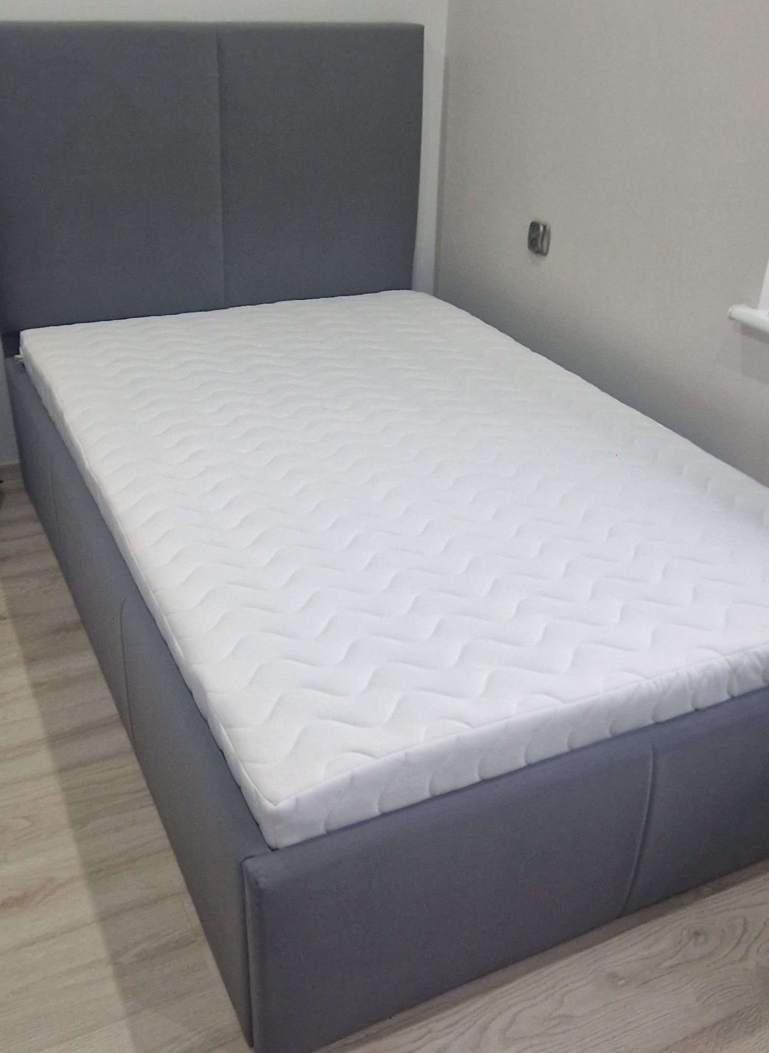 Szare tapicerowane łóżko z materacem w Gostyniu, z widocznym zagłówkiem i bokami, sfotografowane w pomieszczeniu o jasnych ścianach.