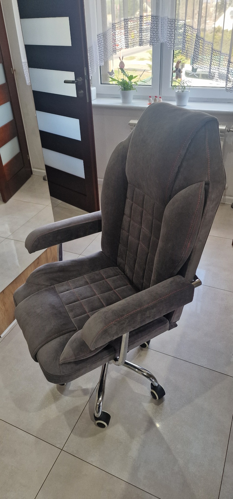 Ergonomiczne krzesło biurowe obite szarym welurem z pikowaniem w romby i czerwonymi przeszyciami, na chromowanej podstawie z kółkami.
