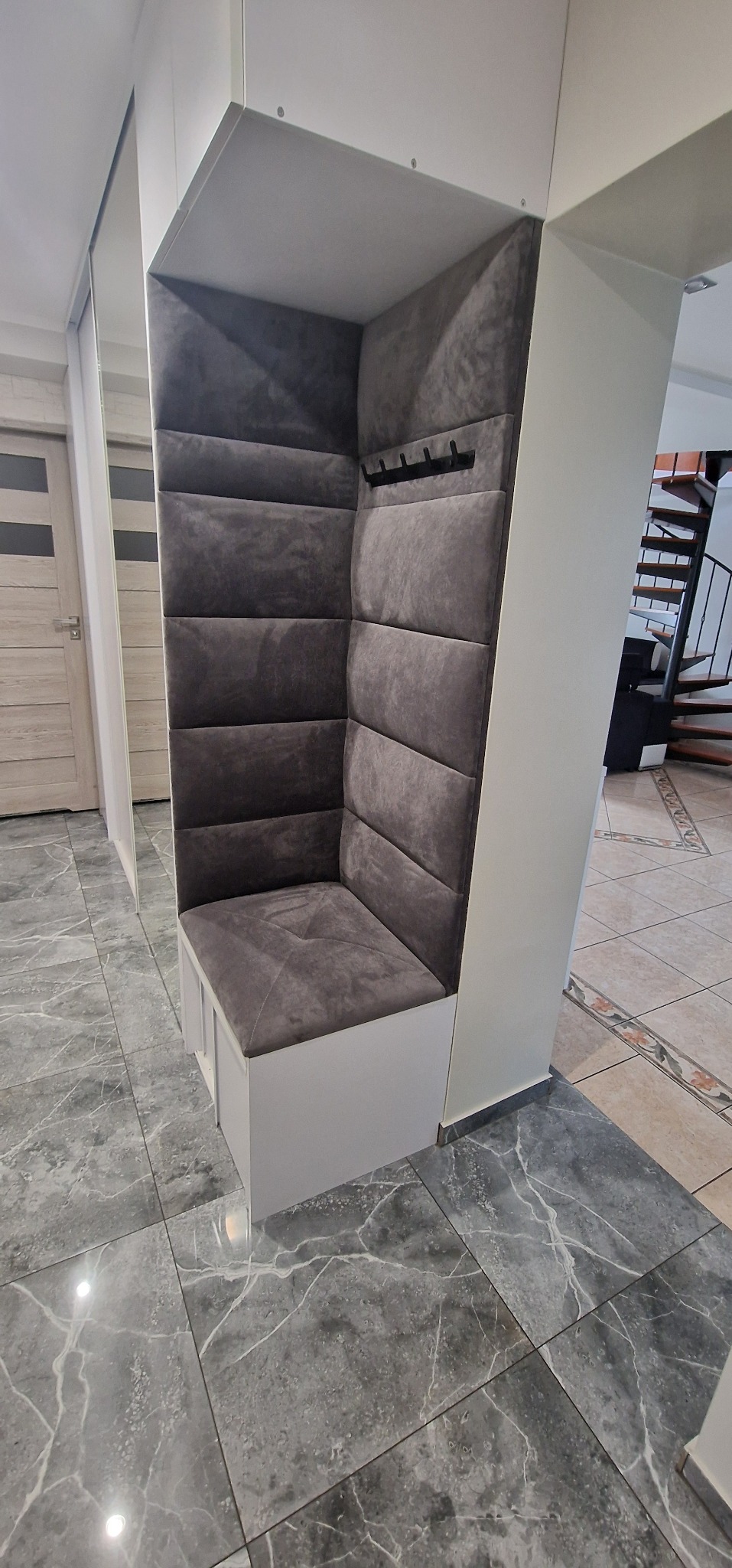 Szara, tapicerowana wnęka z siedziskiem i wieszakami w przedpokoju, element zabudowy na wymiar.