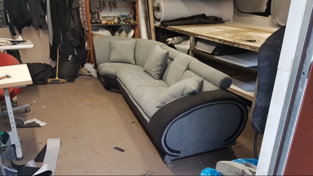 Szary narożnik z poduszkami w warsztacie tapicerskim, widoczne narzędzia i materiały, niedbale ułożone na półkach w tle.