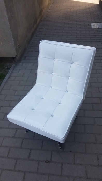 Biały, tapicerowany fotel w stylu modernistycznym z chromowanymi nogami, sfotografowany na brukowanej ścieżce z betonowym murem w tle.