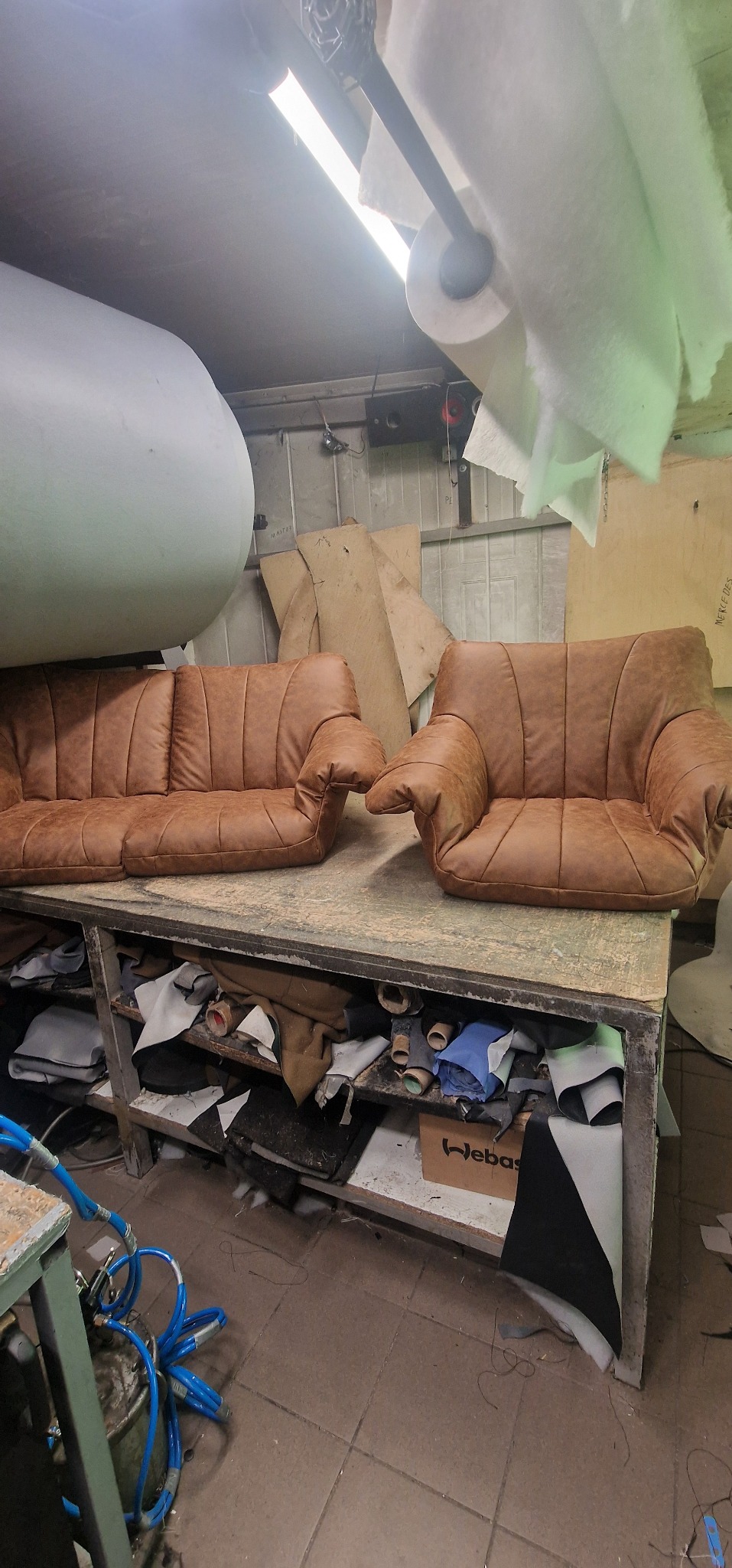 Brązowy fotel i sofa z nową tapicerką na metalowym stole w warsztacie, widoczne materiały i narzędzia na półkach pod stołem.