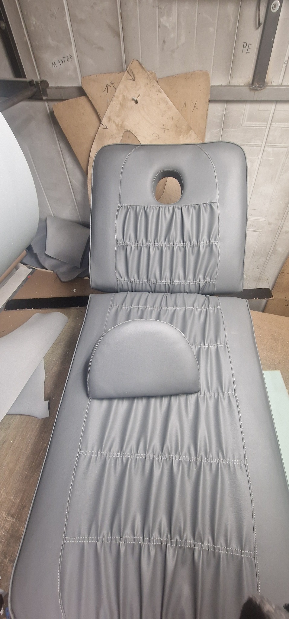 Szare, skórzane siedzisko i oparcie fotela kosmetycznego z otworem na twarz i półokrągłym zagłówkiem, widoczne wzory i szwy, na tle warsztatu.