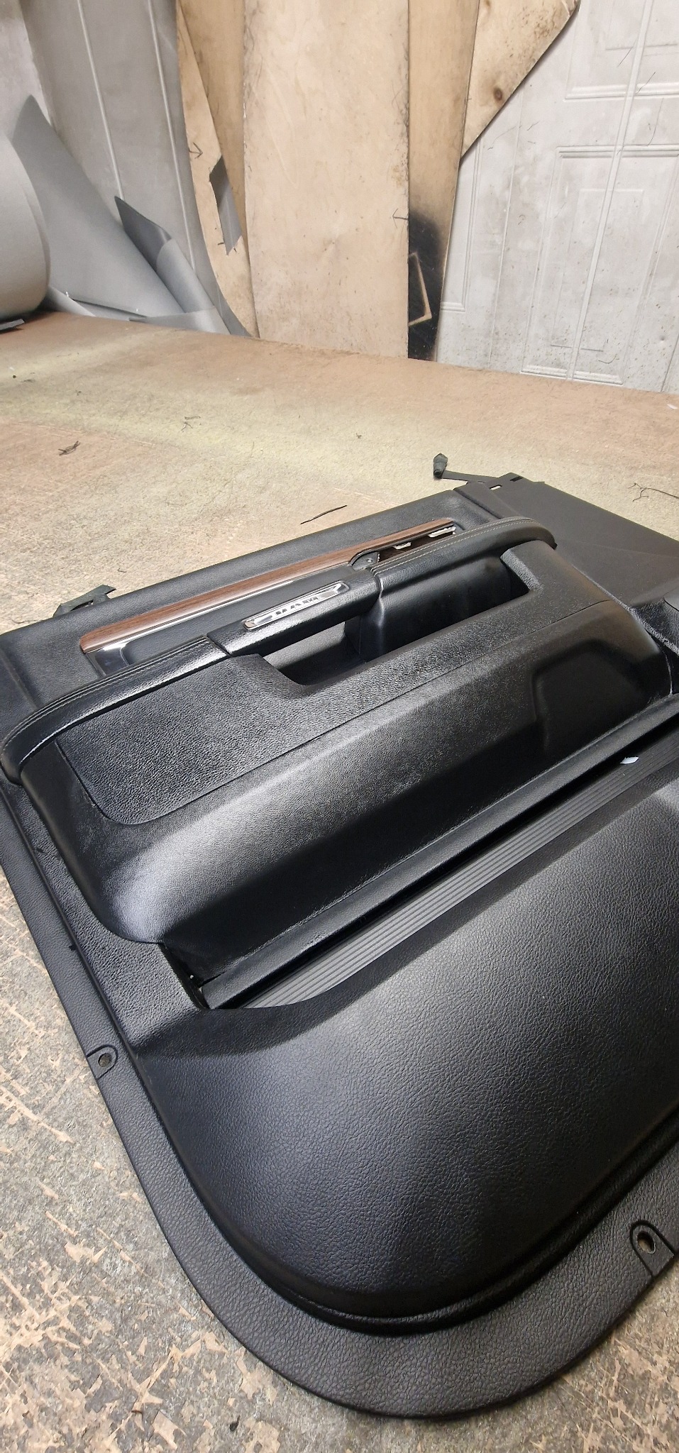 Tapicerowany, czarny panel drzwi samochodowych z drewnianym akcentem, leżący na szorstkiej powierzchni w warsztacie.