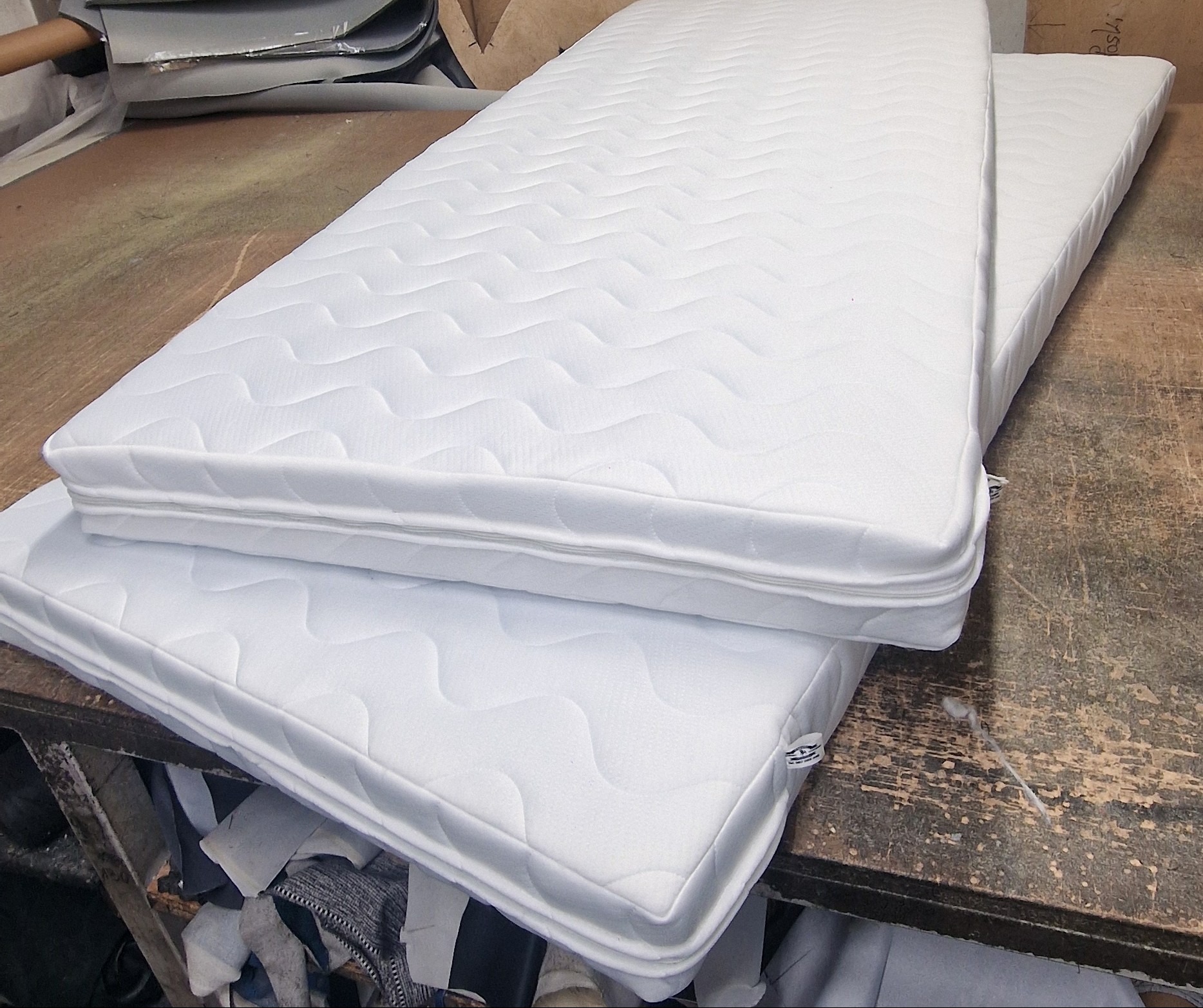 Dwa białe materace z pikowaną powierzchnią i zamkiem błyskawicznym na krawędzi, ułożone jeden na drugim na roboczym stole z widocznymi śladami użytkowania.