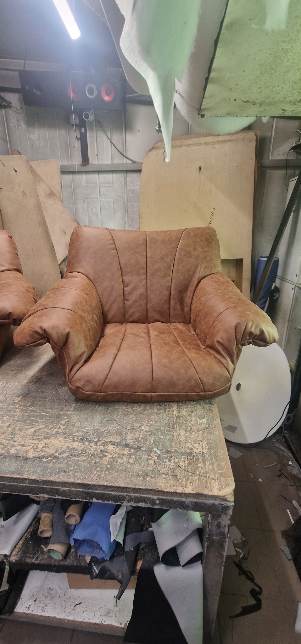 Brązowy fotel tapicerowany na warsztatowym stole z widocznymi materiałami i narzędziami, oświetlenie warsztatowe w tle.