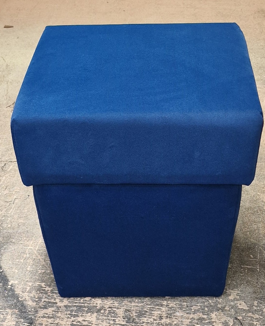 Niebieski, tapicerowany puf w kształcie sześcianu, stojący na szarej, popękanej podłodze.