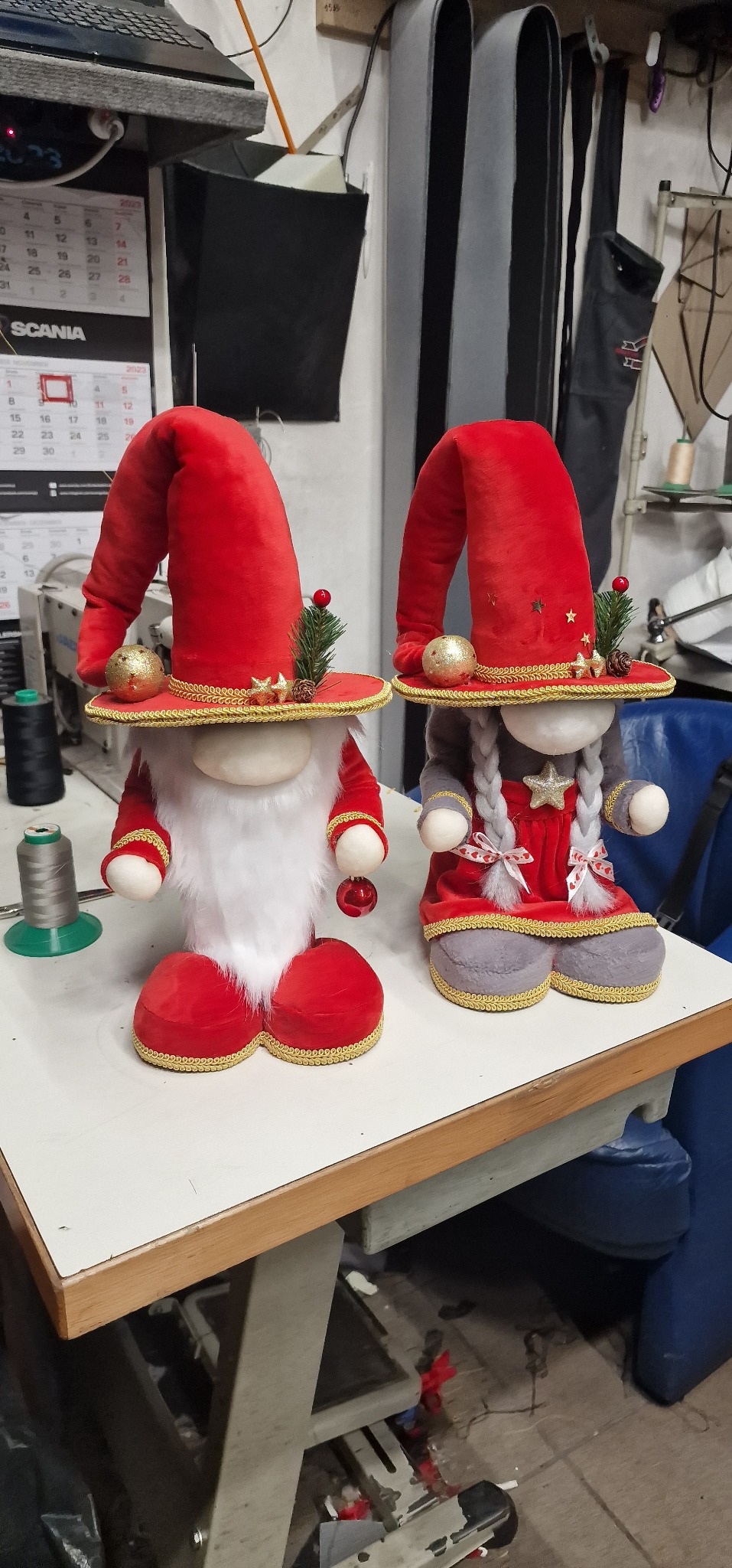 Dwa ręcznie robione, świąteczne krasnale z filcu w czerwono-szarych kolorach, stojące na stole w pracowni, widoczne nici i kalendarz w tle.