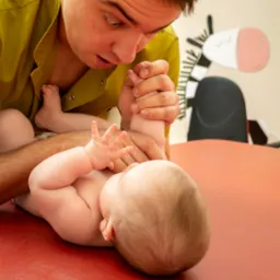 Mężczyzna w zielonej koszuli bada odruchy niemowlęcia leżącego na czerwonym stole, w tle naklejka zebry na ścianie.