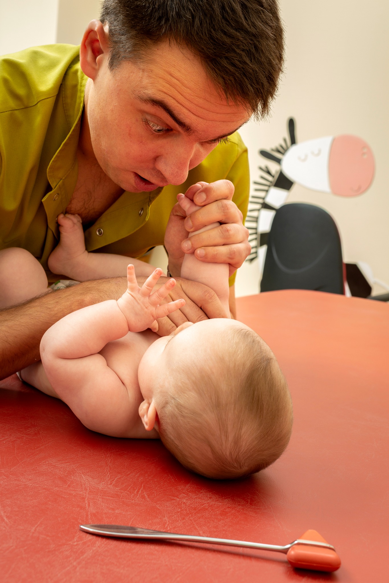 Mężczyzna w zielonej koszuli bada odruchy niemowlęcia leżącego na czerwonym stole, w tle naklejka zebry na ścianie.