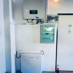 Elektrowniesloneczne.tech - Wnętrze pomieszczenia technicznego z zamontowanym inwerterem, akumulatorem energii, rozdzielnicami elektrycznymi i gaśnicami w tle, widoczne naklejki ostrzegawcze.