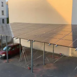 Zadaszenie parkingu z panelami słonecznymi, czerwony samochód pod spodem, drabina oparta o konstrukcję, budynek w tle.