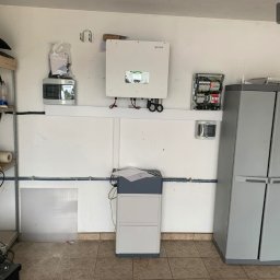Elektrowniesloneczne.tech - Wnętrze garażu z zamontowanym inwerterem firmy Sofar, rozdzielnicą elektryczną, magazynem energii na podłodze oraz metalowym regałem z narzędziami i akcesoriami.