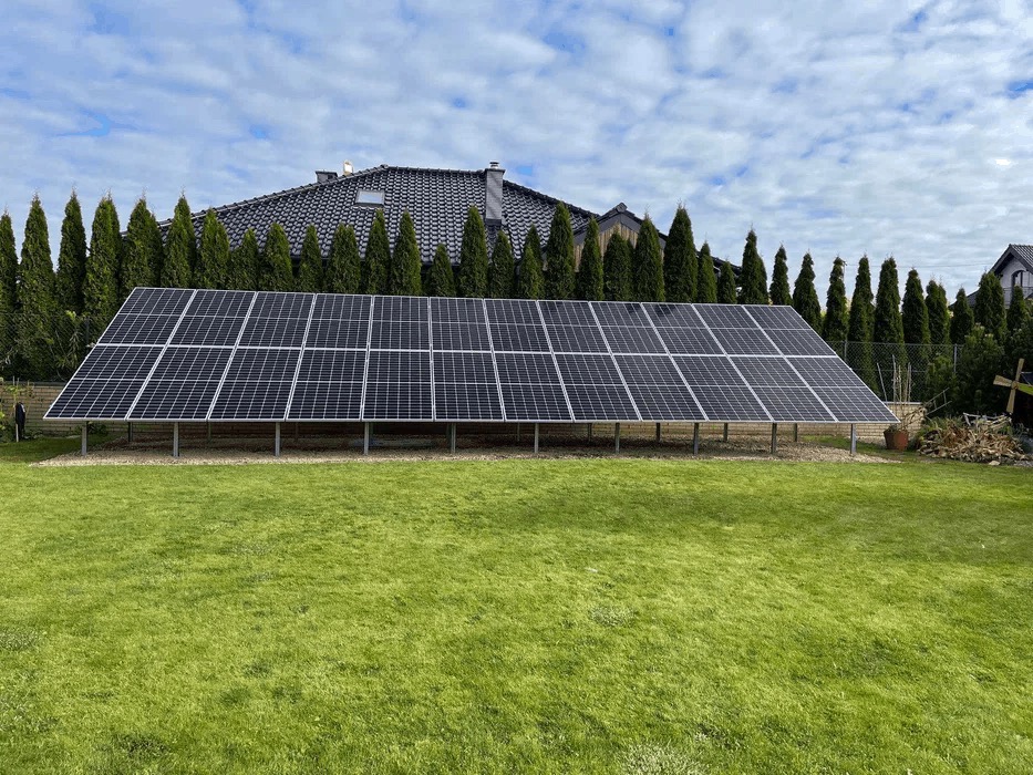 Rząd paneli słonecznych ustawionych na metalowych nóżkach na trawniku, z rzędem wysokich, smukłych drzew iglastych i domem w tle, pod błękitnym niebem z chmurami.