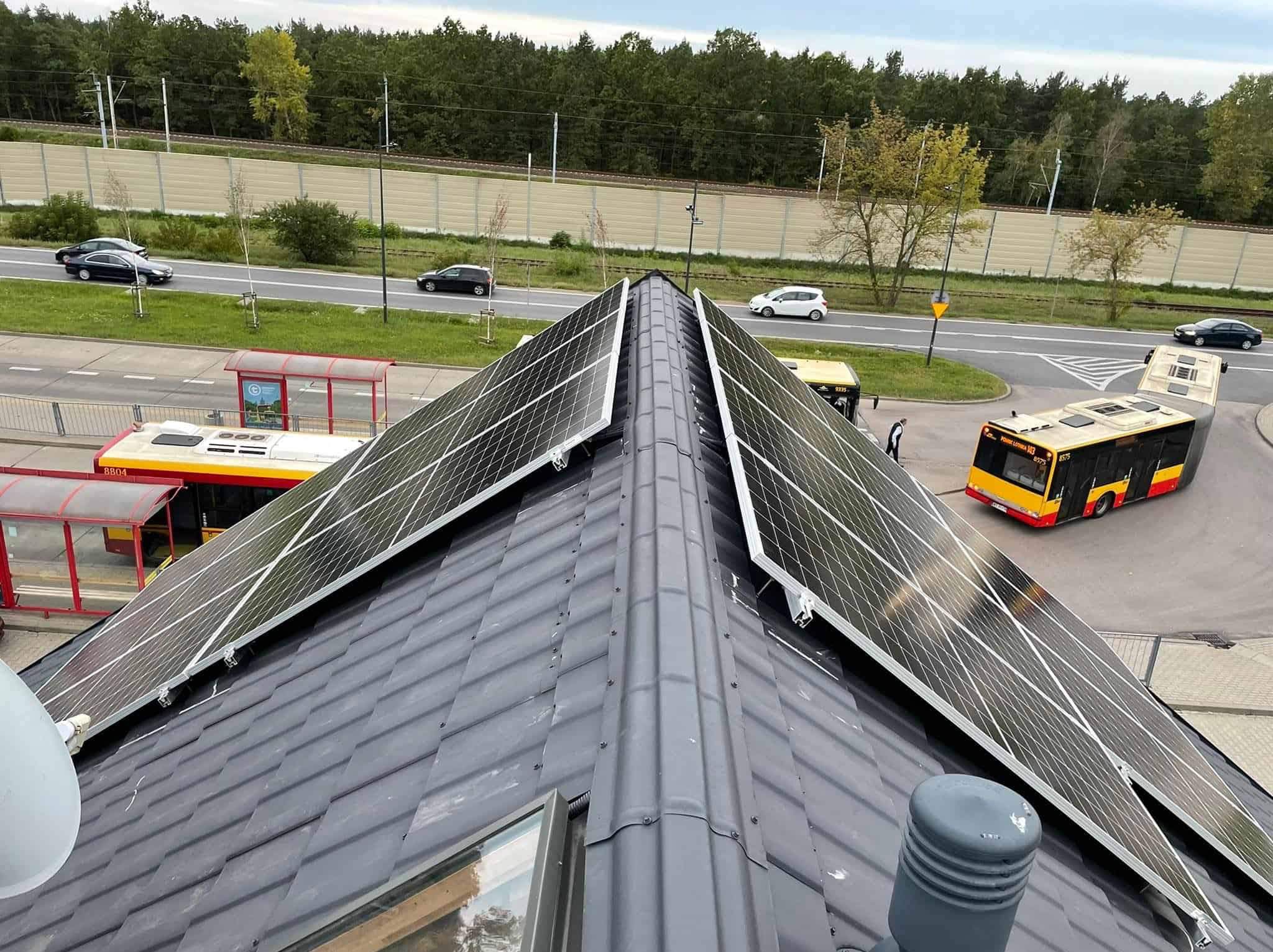 Panele słoneczne na dachu budynku z widokiem na przystanek autobusowy i ruchliwą ulicę.