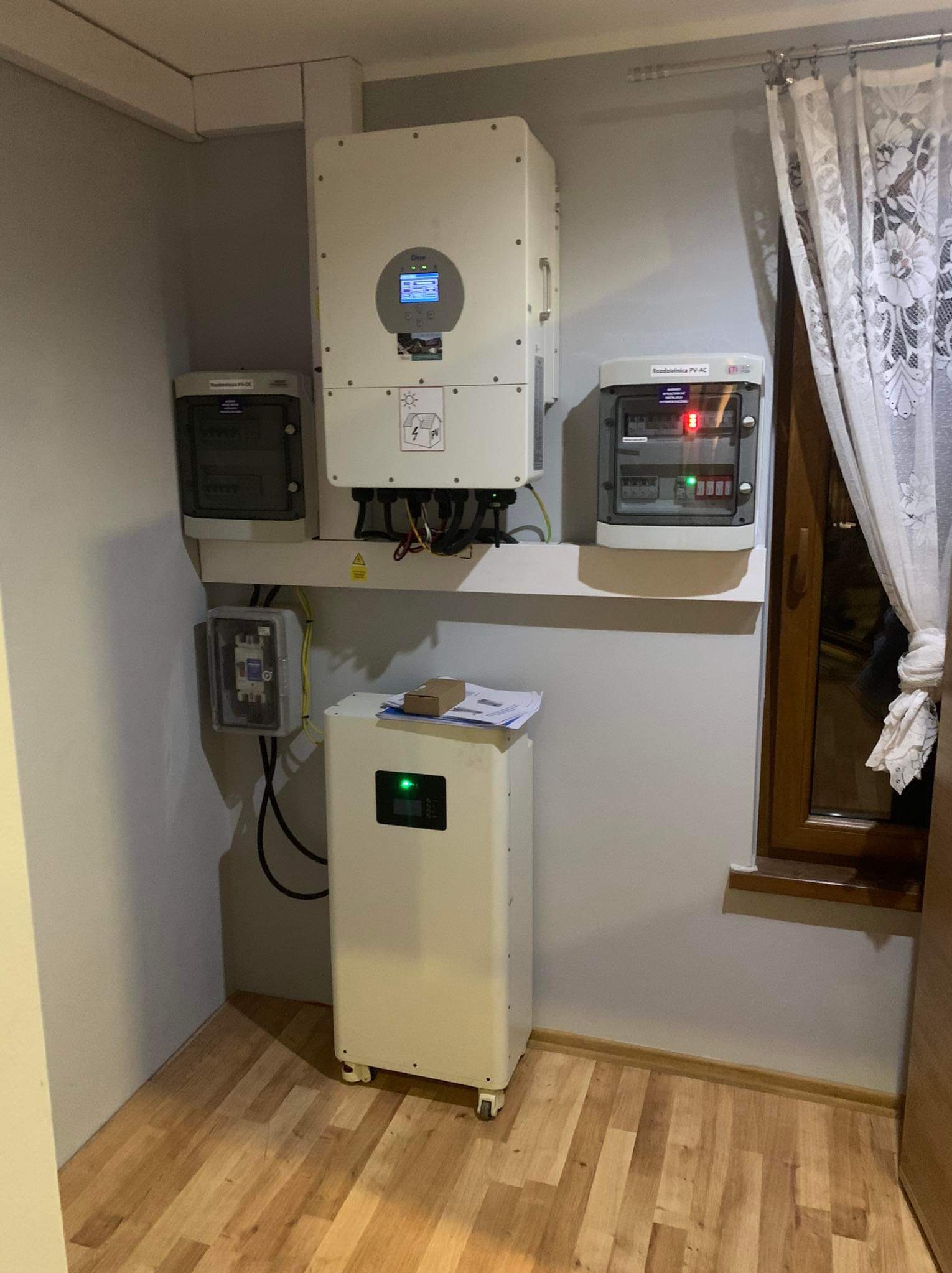 Wnętrze pomieszczenia z zainstalowanym systemem fotowoltaicznym: falownik z wyświetlaczem, rozdzielnice AC i DC, oraz magazyn energii z zieloną diodą LED, na tle drewnianej podłogi i okna z zasłoną.