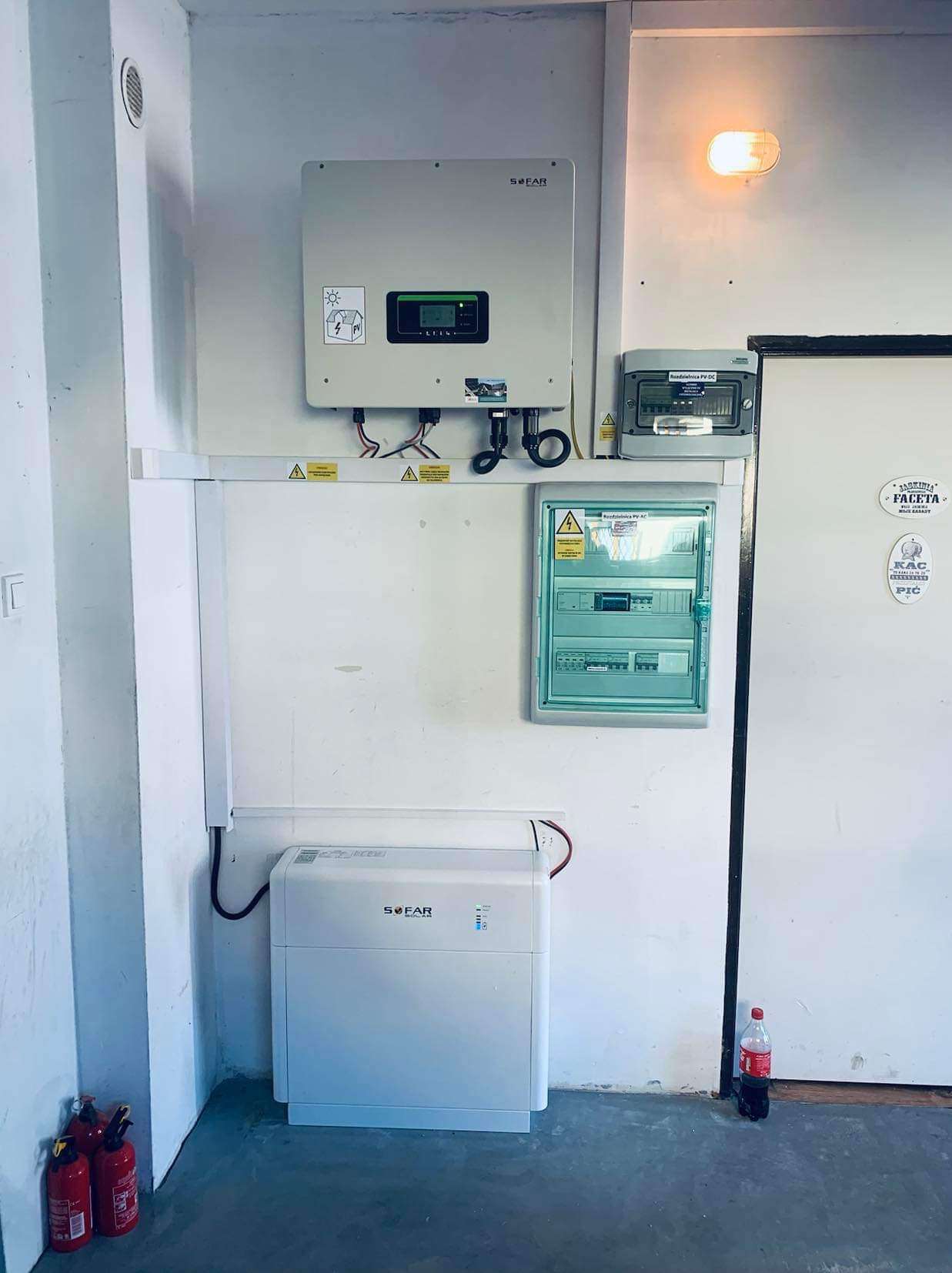 Wnętrze pomieszczenia technicznego z zamontowanym inwerterem, akumulatorem energii, rozdzielnicami elektrycznymi i gaśnicami w tle, widoczne naklejki ostrzegawcze.