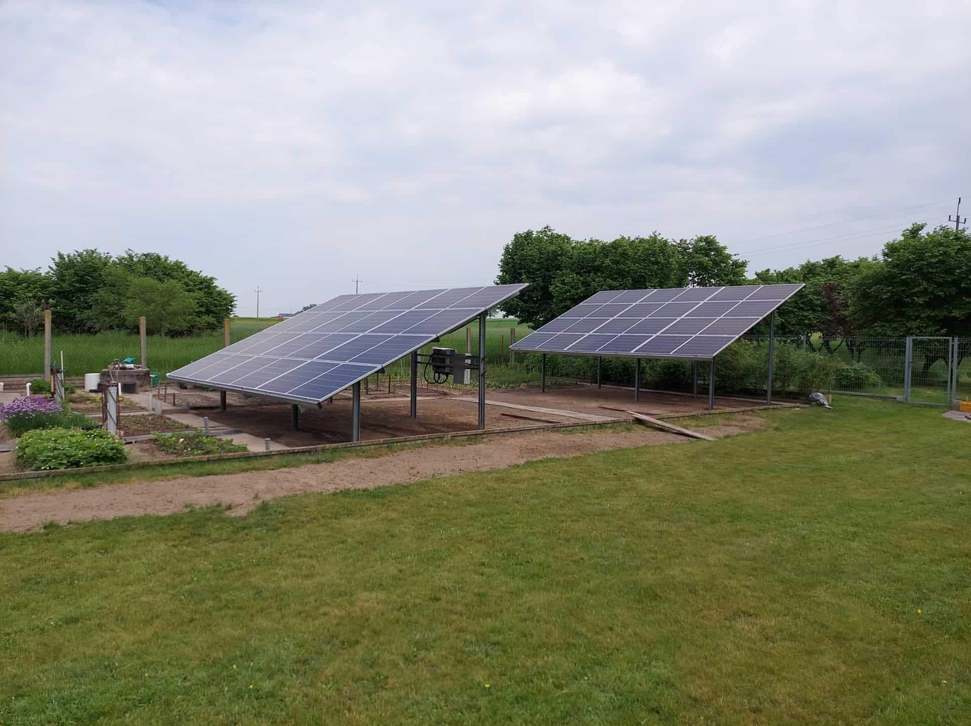 Dwa panele słoneczne na metalowej konstrukcji, zainstalowane na trawniku w ogrodzie, z widocznymi grządkami kwiatowymi i drzewami w tle.