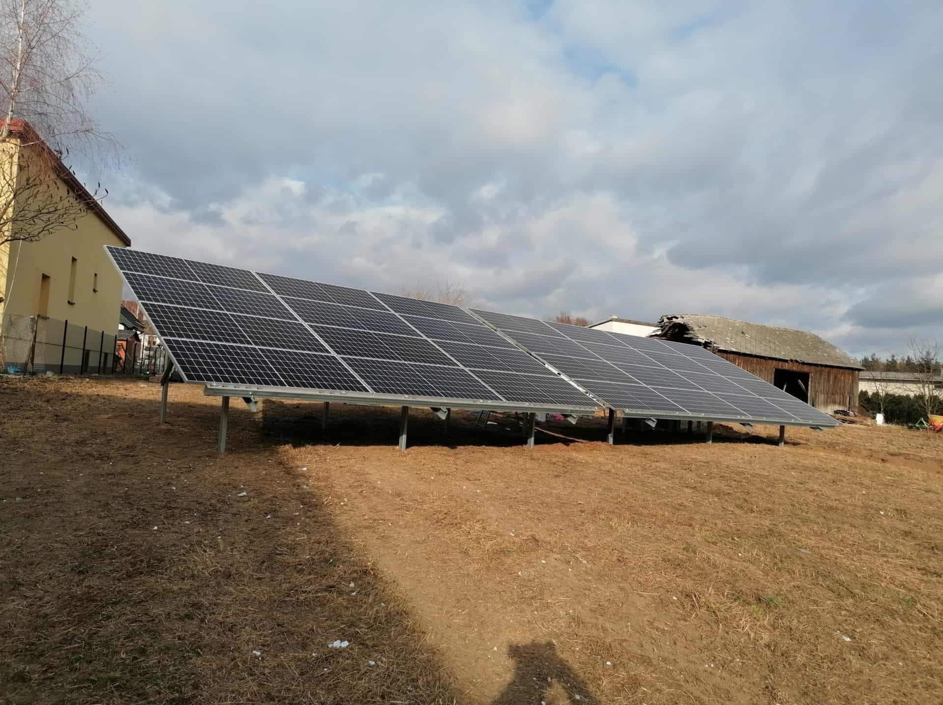 Rząd paneli słonecznych na metalowej konstrukcji, zamontowany na wzgórzu z suchą trawą, w tle budynek mieszkalny i drewniana stodoła pod pochmurnym niebem.