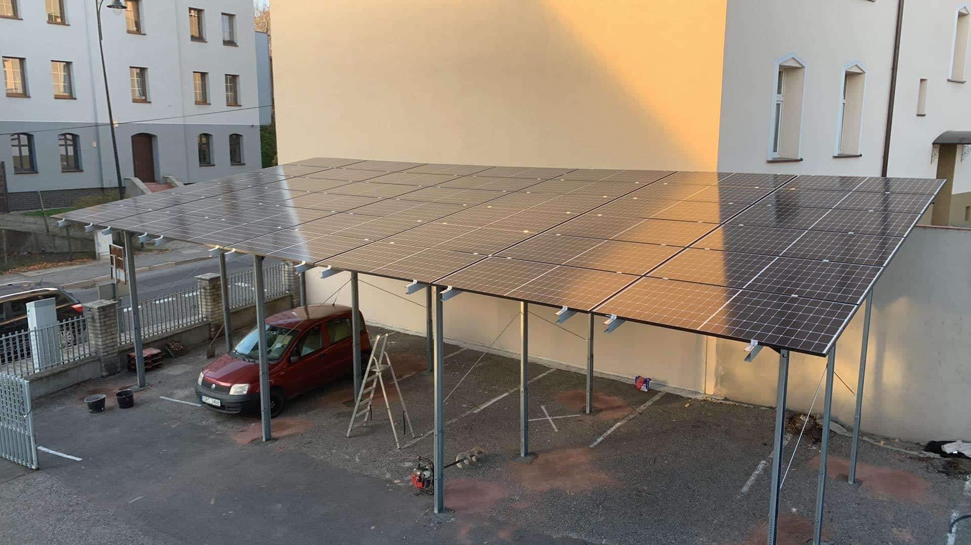 Zadaszenie parkingu z panelami słonecznymi, czerwony samochód pod spodem, drabina oparta o konstrukcję, budynek w tle.