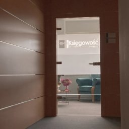 PM SSC SPÓŁKA Z OGRANICZONĄ ODPOWIEDZIALNOŚCIĄ - Widok z korytarza na przeszklone drzwi z napisem 'Księgowość' i logo, za którymi widać wnętrze biura z turkusowym fotelem i kwiatami na stoliku.