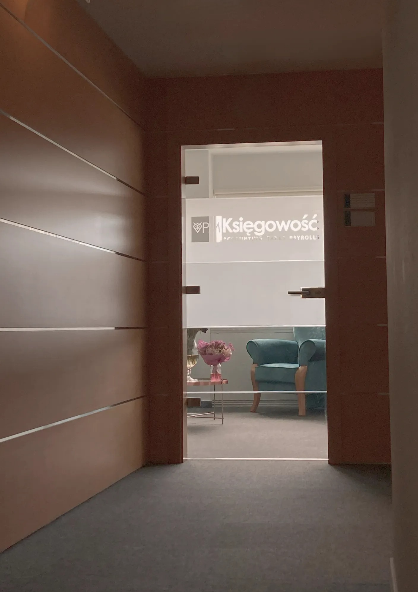 Widok z korytarza na przeszklone drzwi z napisem 'Księgowość' i logo, za którymi widać wnętrze biura z turkusowym fotelem i kwiatami na stoliku.