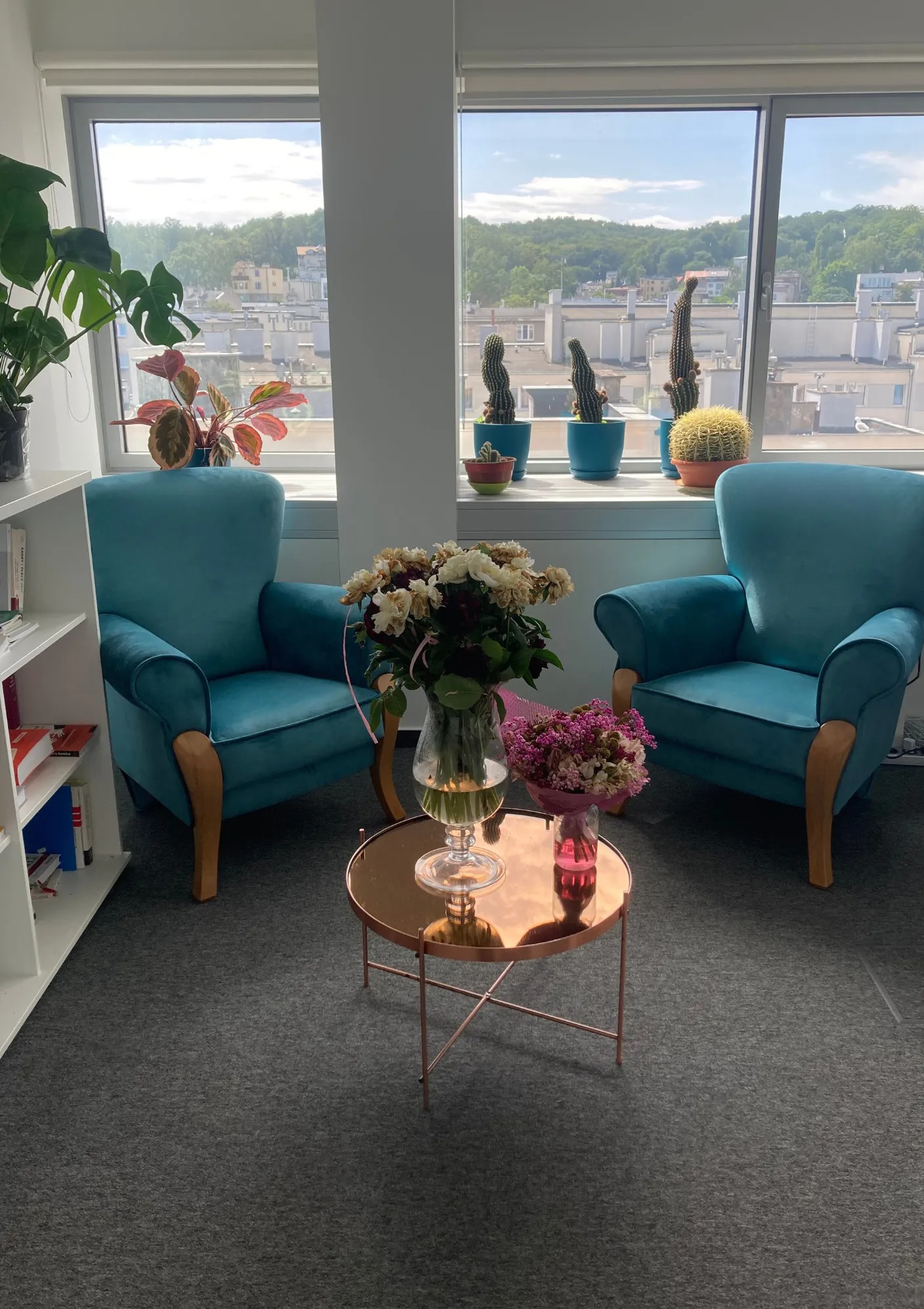 Jasne wnętrze biura z dwoma turkusowymi fotelami, stolikiem kawowym z bukietami kwiatów, widokiem na miasto zza okna, dekoracjami roślinnymi i regałem z książkami.