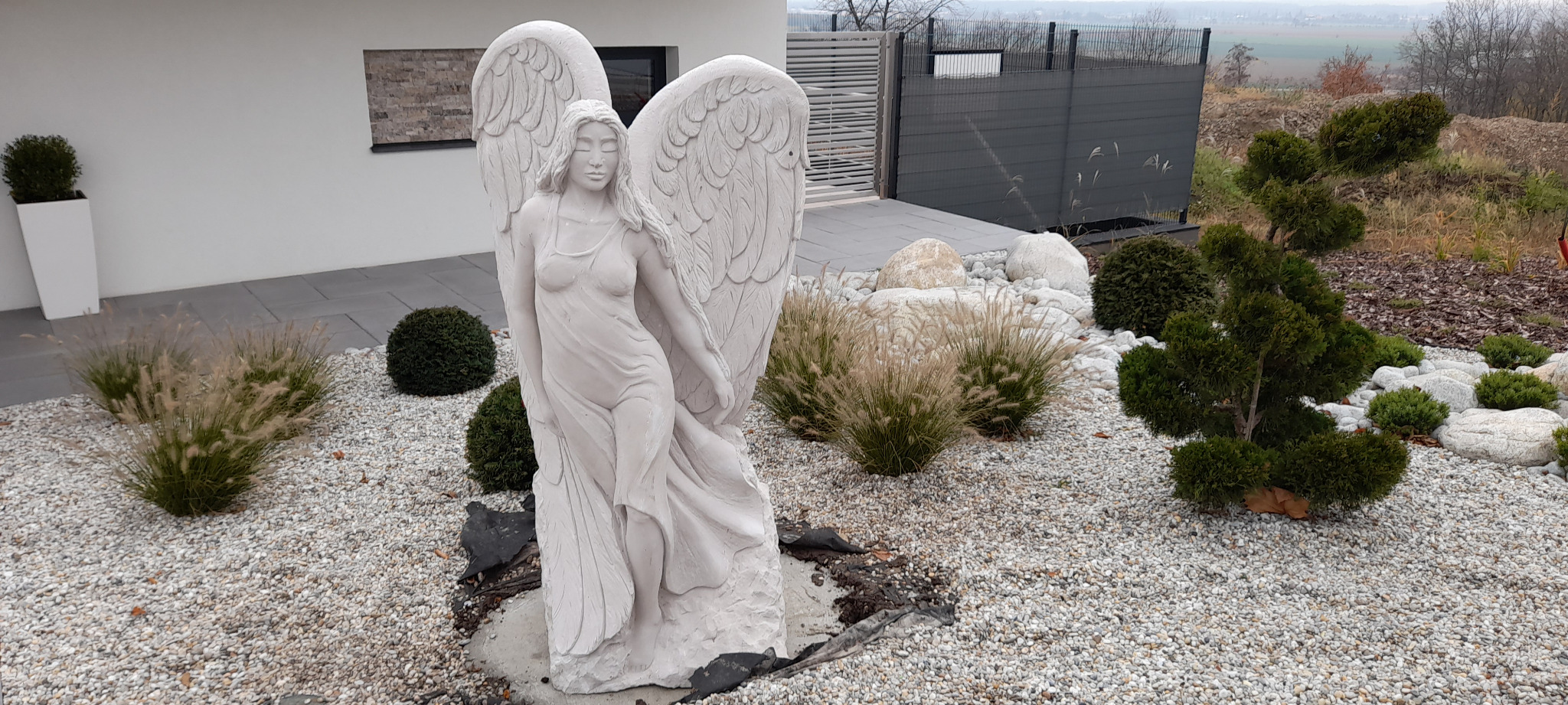 Ogród z rzeźbą anioła, ozdobnymi krzewami i kamieniami. Nowoczesny dom z szarą elewacją w tle. Dekoracyjny element krajobrazu.