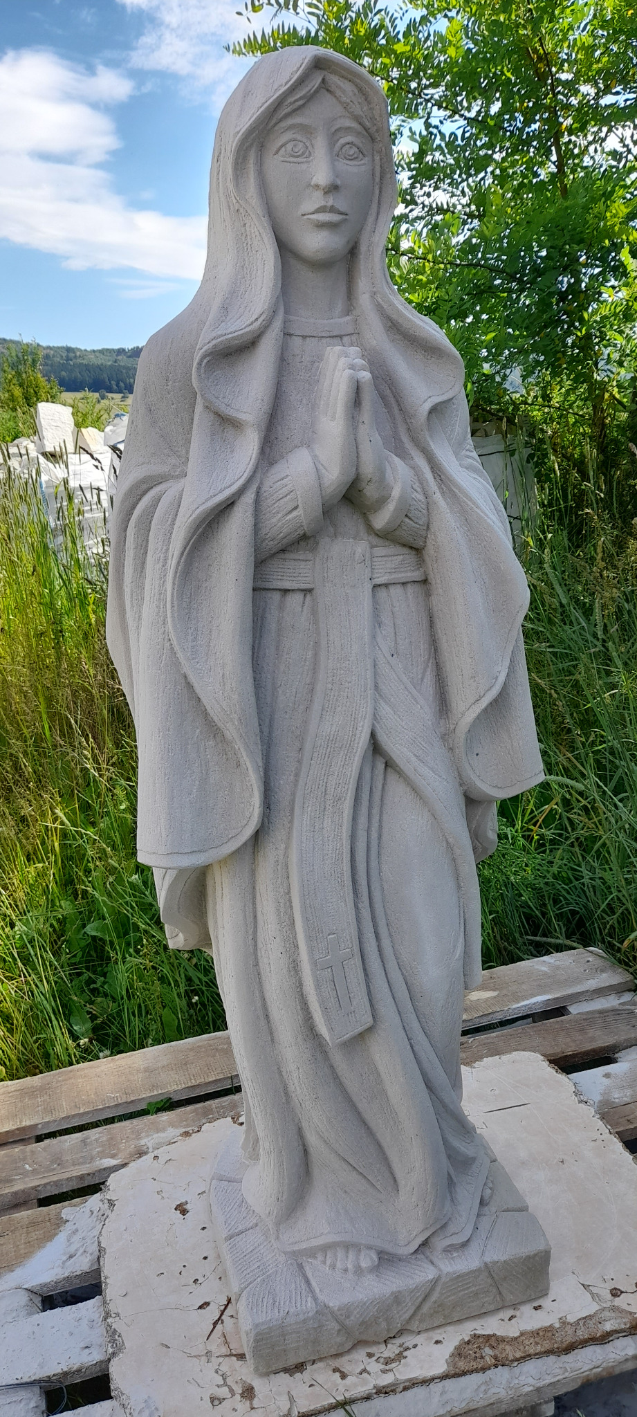 Statua Matki Boskiej z betonu architektonicznego, z detalami krzyża na szarfie, na tle zieleni i błękitnego nieba. Ręcznie wykonana rzeźba ogrodowa.