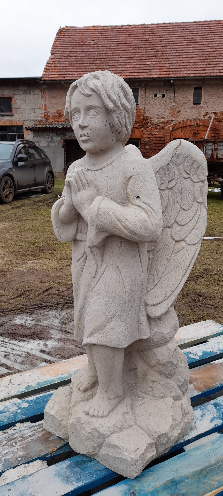 Betonowa rzeźba anioła z rozłożonymi skrzydłami na tle ceglanego budynku i szarego samochodu. Anioł ma złożone ręce do modlitwy.