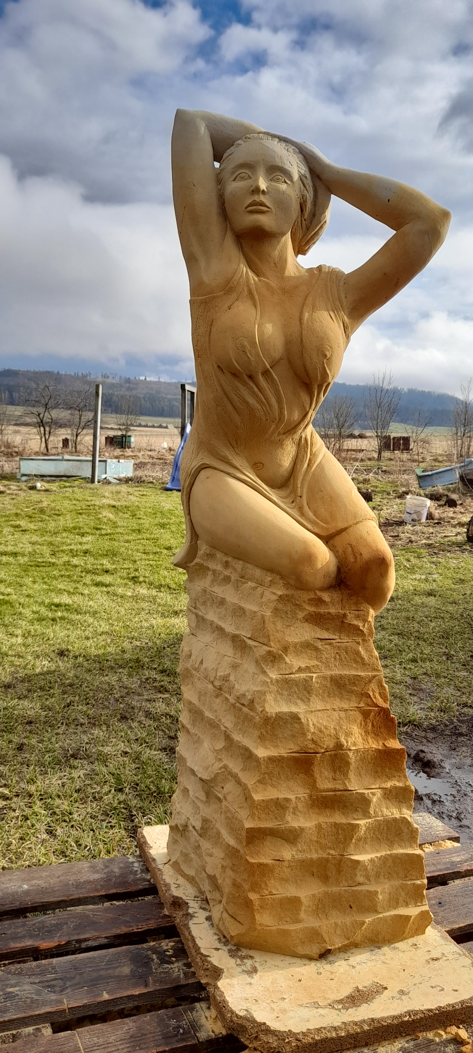 Piaskowa rzeźba kobiety w plenerze, na drewnianej palecie. W tle łąka i wzgórza pod pochmurnym niebem. Artystyczne detale i naturalne światło podkreślają unikalność dzieła.