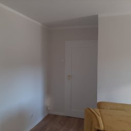 Met -Bud - Jasne, świeżo pomalowane ściany w pokoju z białą listwą przysufitową i drzwiami. Fragment żółtej sofy w prawym dolnym rogu.